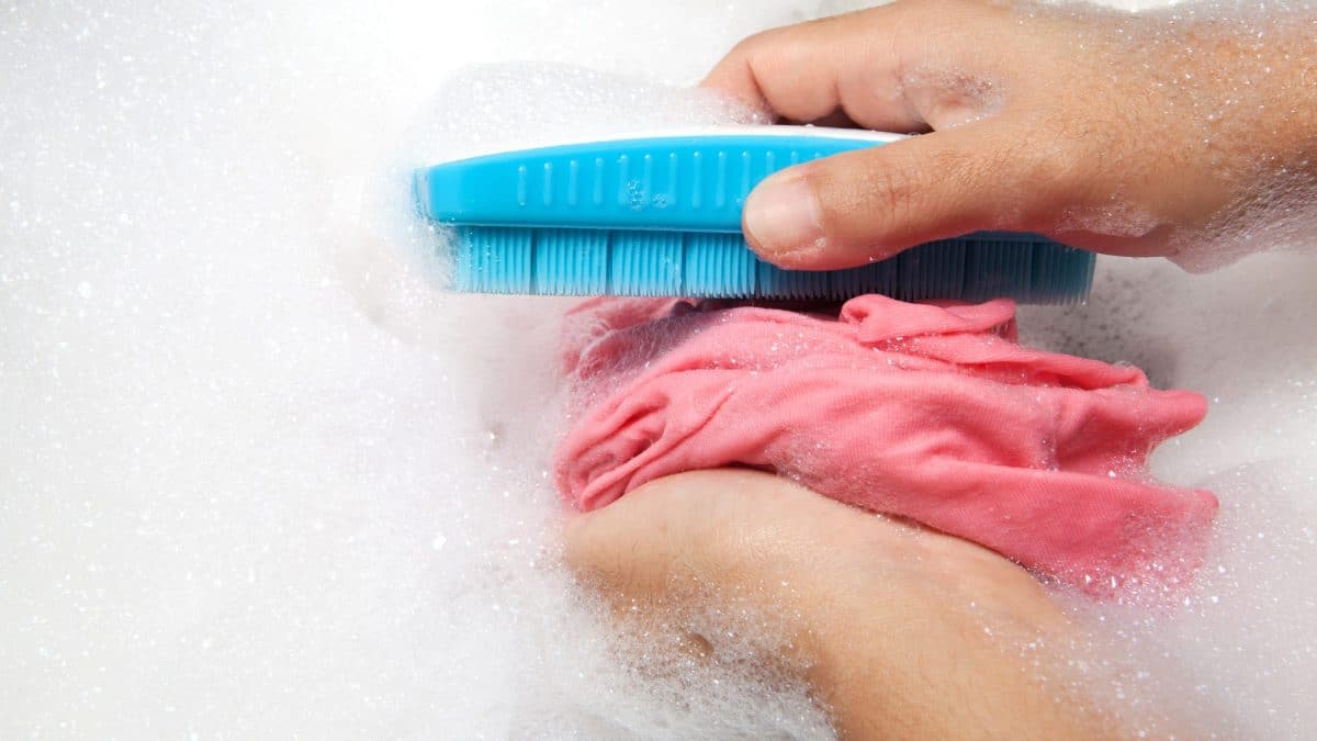 A hand works a soft blue brush across pink fabric in a basin of suds