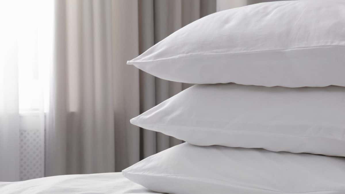 Three white pillows stacked on a made bed beside a curtained bedroom window
