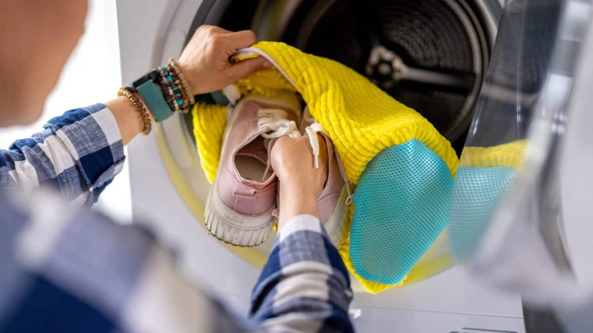 Hands tucking pink sneakers inside a yellow mesh laundry bag at the washer drum