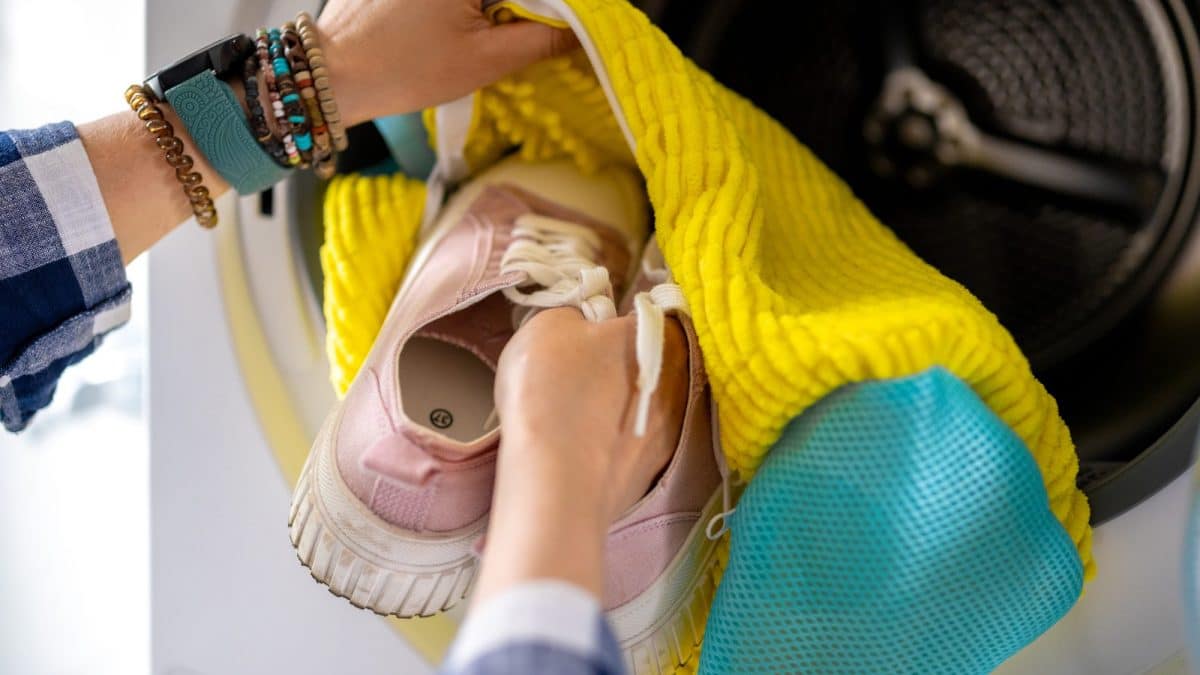 Hands lowering a pair of fabric sneakers inside a mesh laundry bag into a washing machine