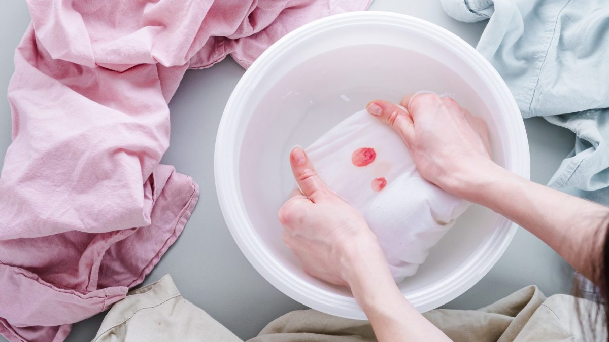Hands pressing a blood-spotted white garment into a white basin filled with cold water
