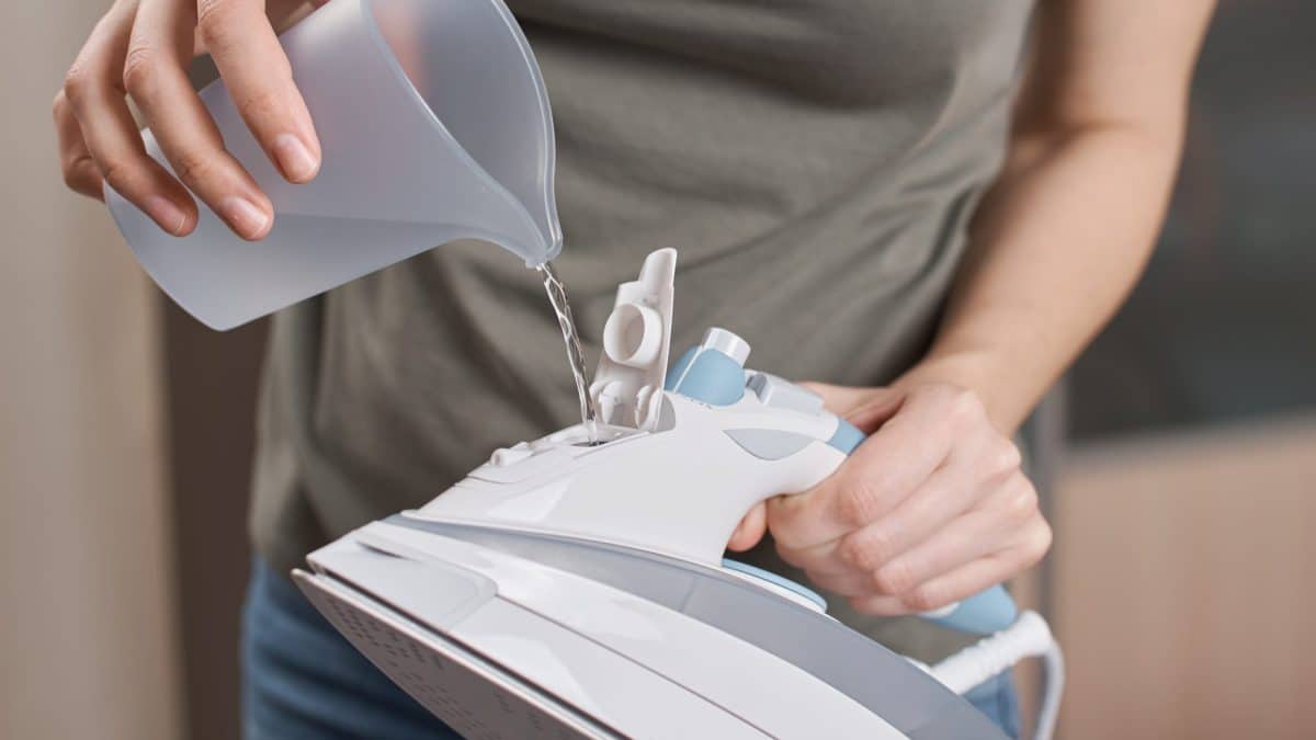 Hand pouring water from a grey jug into the fill opening of a steam iron