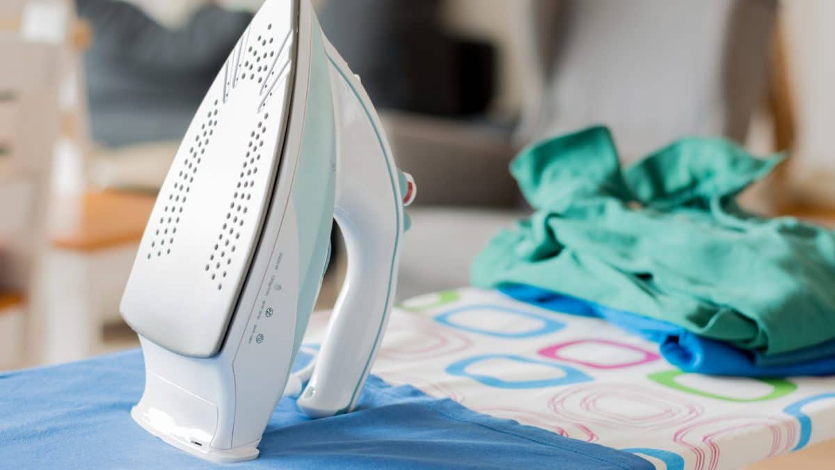 White steam iron standing upright on a blue ironing board beside folded laundry indoors