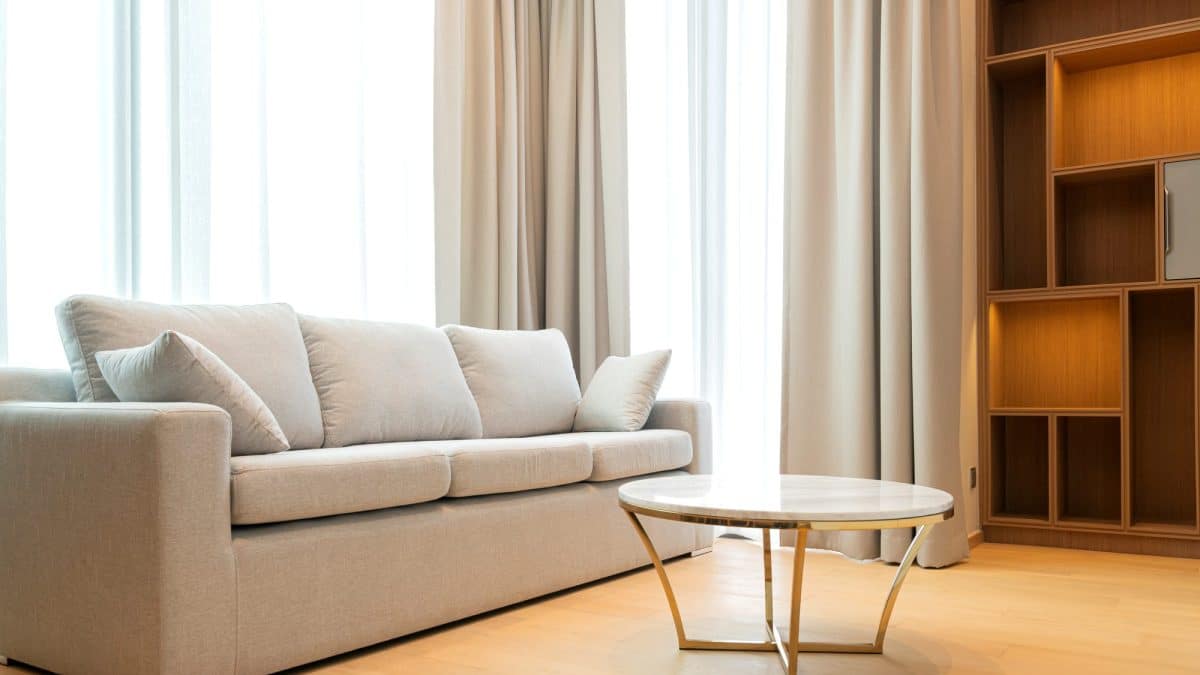 Beige fabric sofa with cushions beside a round marble table in a sunlit living room