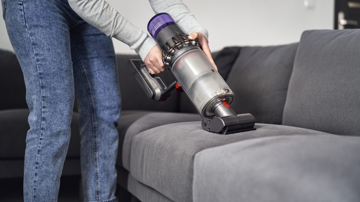 Woman running a cordless vacuum nozzle across the seat cushion of a grey fabric sofa