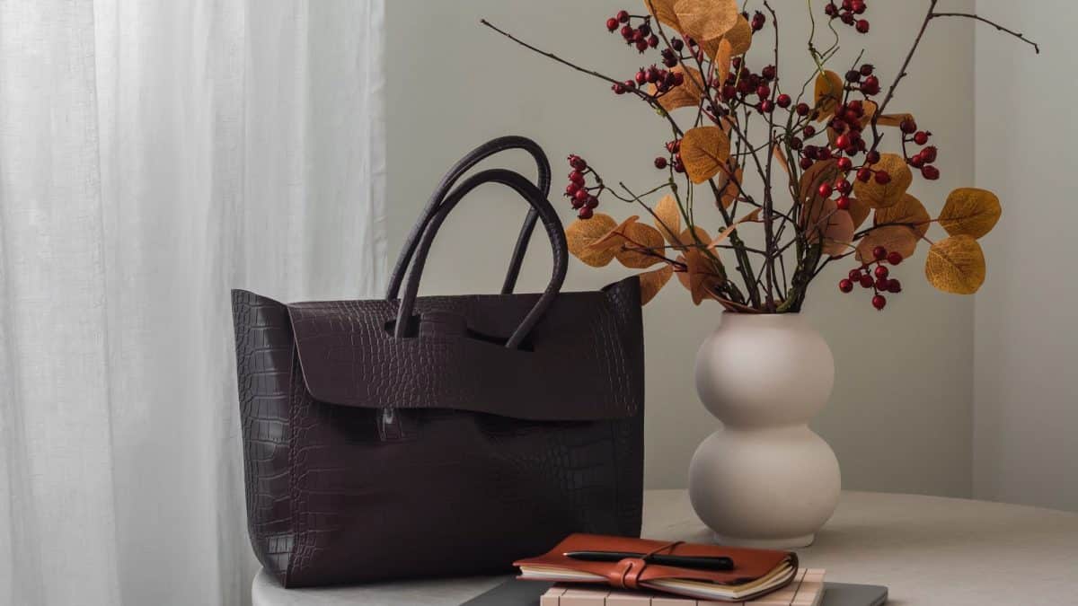 Dark brown structured leather handbag standing on a cloth-covered side table beside dried flowers