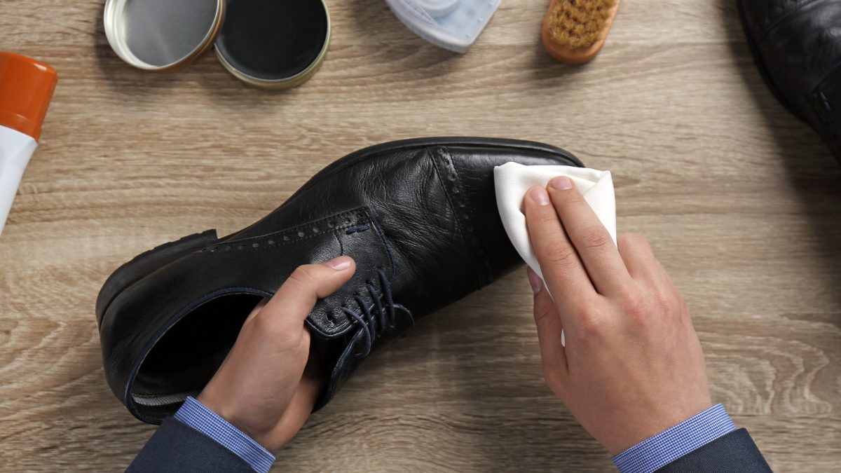 Hands wiping black leather with a soft white cloth beside tins of conditioner and a brush