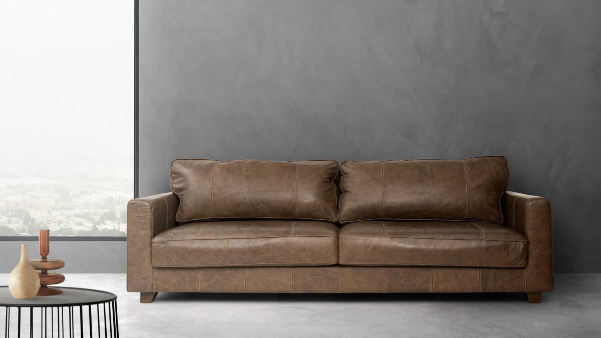 Brown leather three-seat sofa against a grey concrete wall beside a low round table