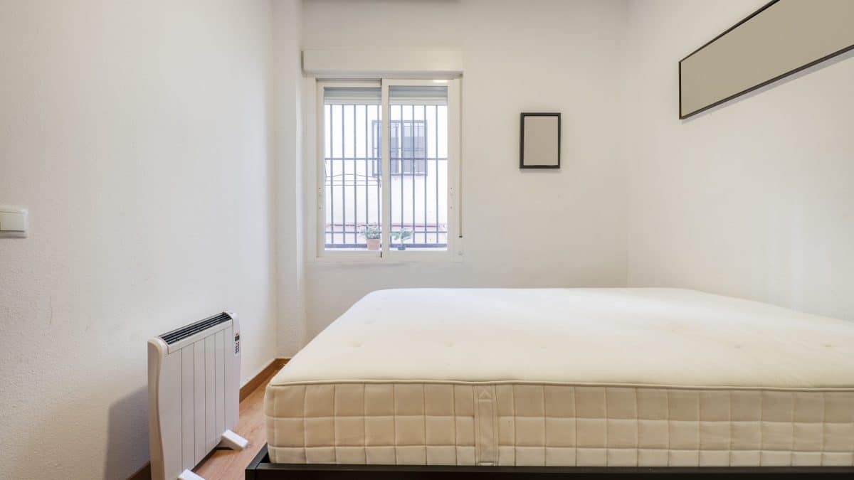 Bare quilted mattress on a wooden bed frame in a plain bedroom by a window
