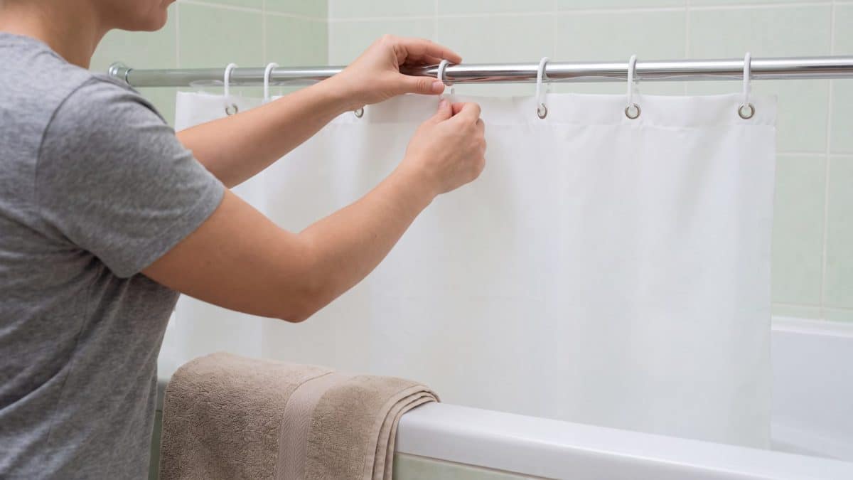 Hands threading a white shower curtain onto metal rings on a bathroom rod