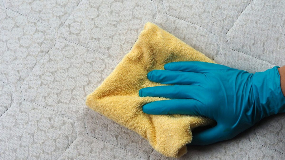 Gloved hand pressing a folded yellow microfibre cloth flat onto the quilted surface of a mattress