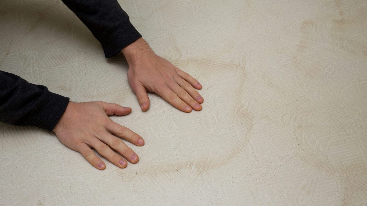 Hands on a mattress beside a large dried yellow-brown stain