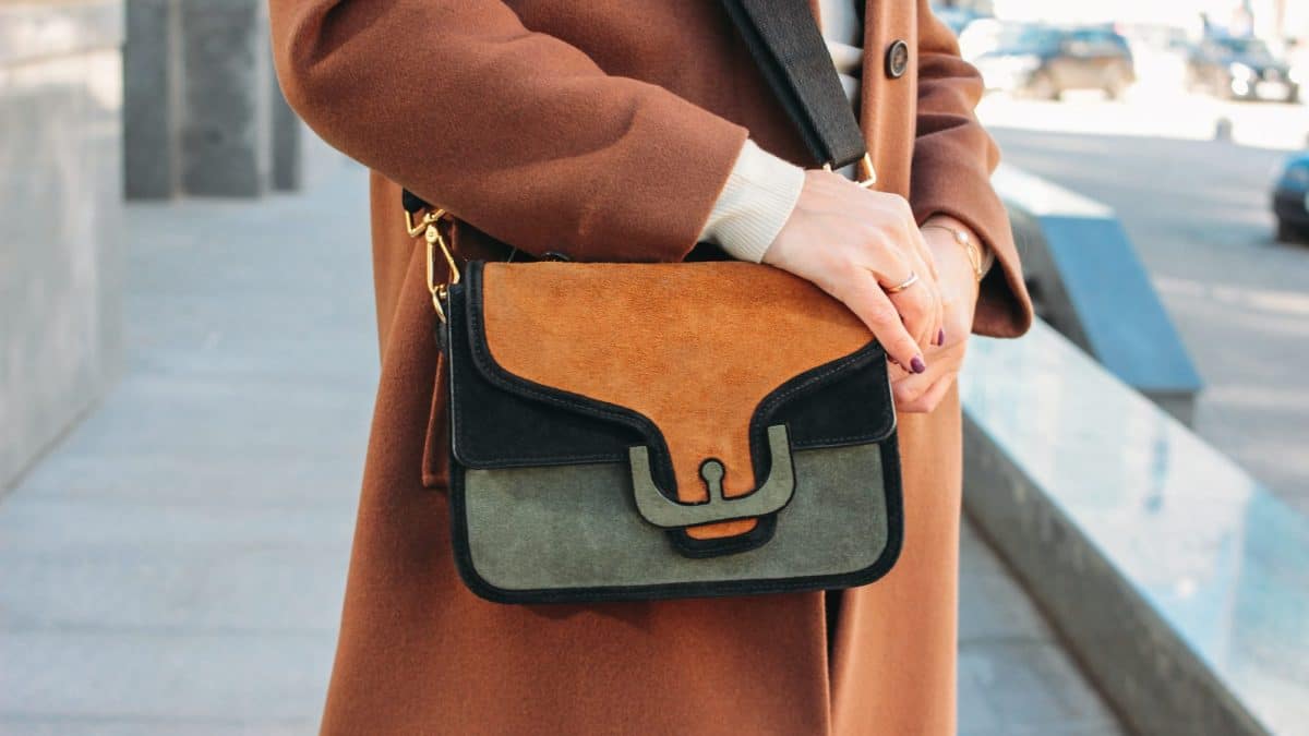 Colour-blocked suede crossbody bag with metal hardware resting against a wool coat