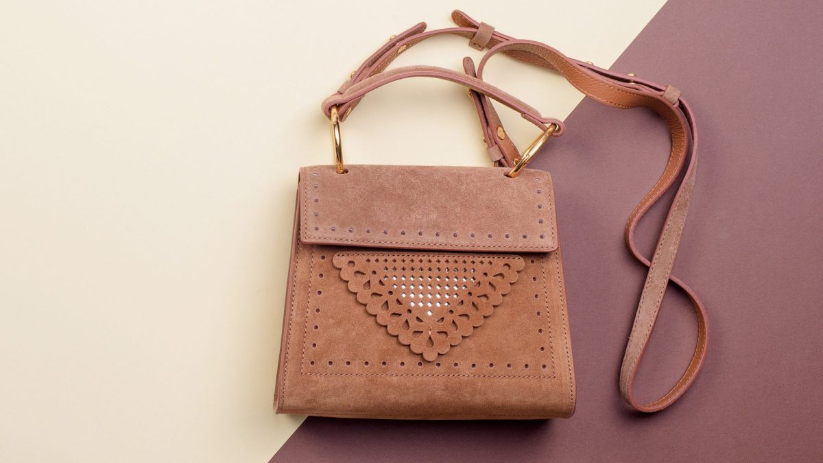 Dusty pink suede crossbody bag with gold hardware and long shoulder strap on two-tone surface