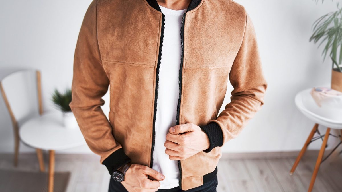 Man in a tan suede bomber jacket standing in a bright domestic room with pale floorboards