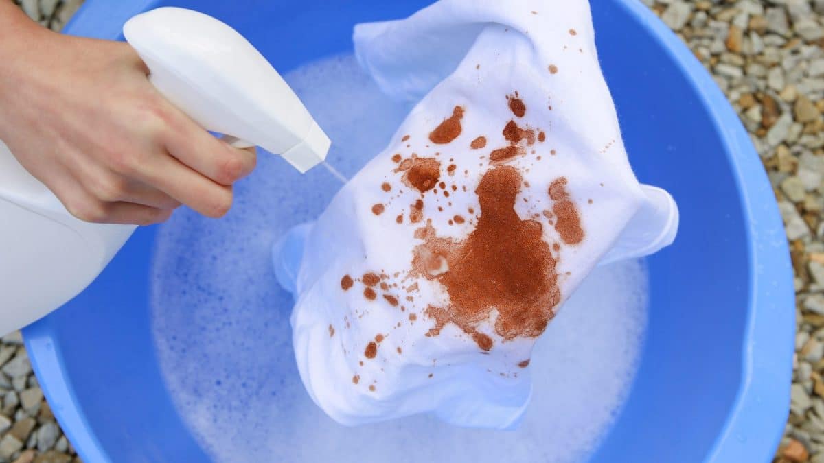 Hands spraying stain remover onto a red-brown stain on white fabric soaking in a basin