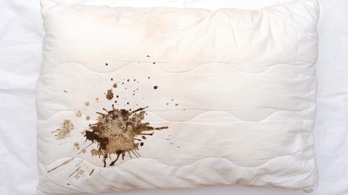 Large brown stain spread across a white pillow lying on crumpled white bed sheets