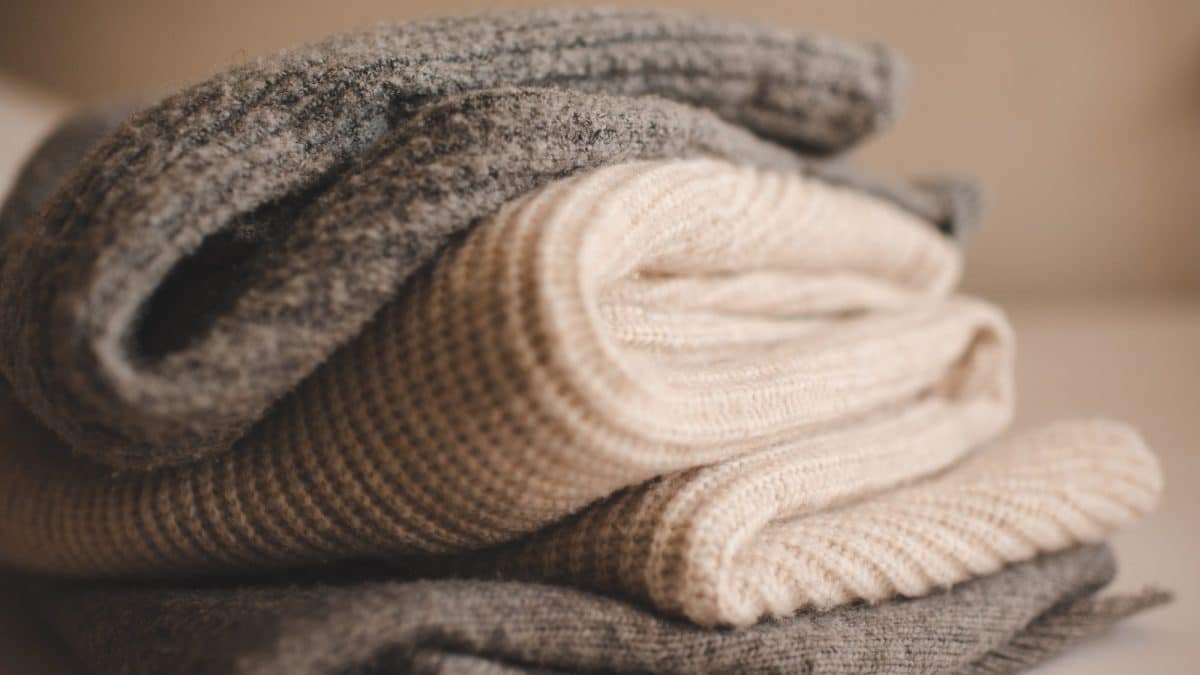 Stack of folded fine-knit sweaters in grey and cream resting on a bed