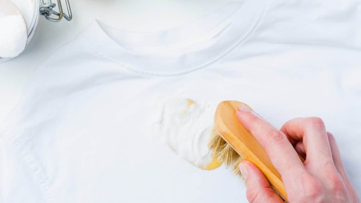 Hand working a baking soda paste into a yellow stain on a white t-shirt with a wooden brush
