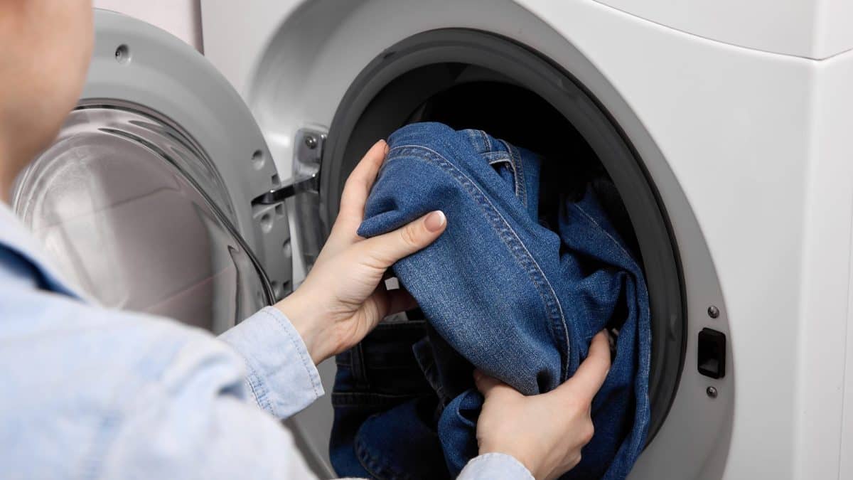 Two hands lowering dark blue jeans into the open drum of a front-loading washing machine
