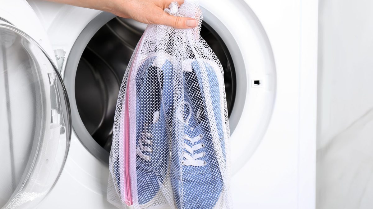 Hand lowering a zipped mesh laundry bag holding two fabric sneakers into a washing machine