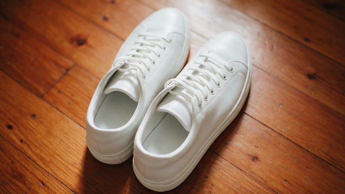 A pair of white leather sneakers with white rubber soles resting on a wooden home floor